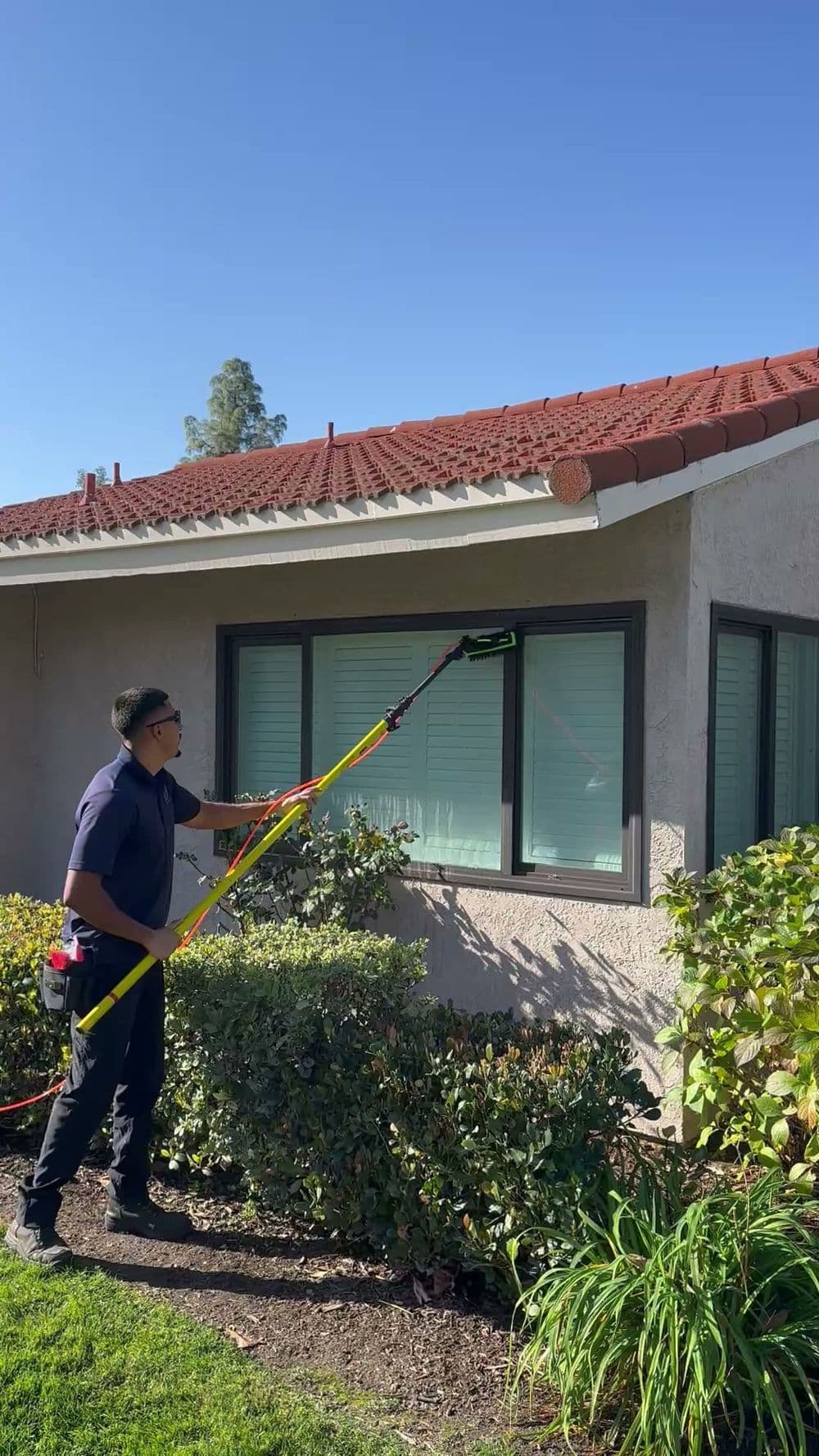 Man using a pole cleaner to wash windows on a house exterior under clear blue skies.
