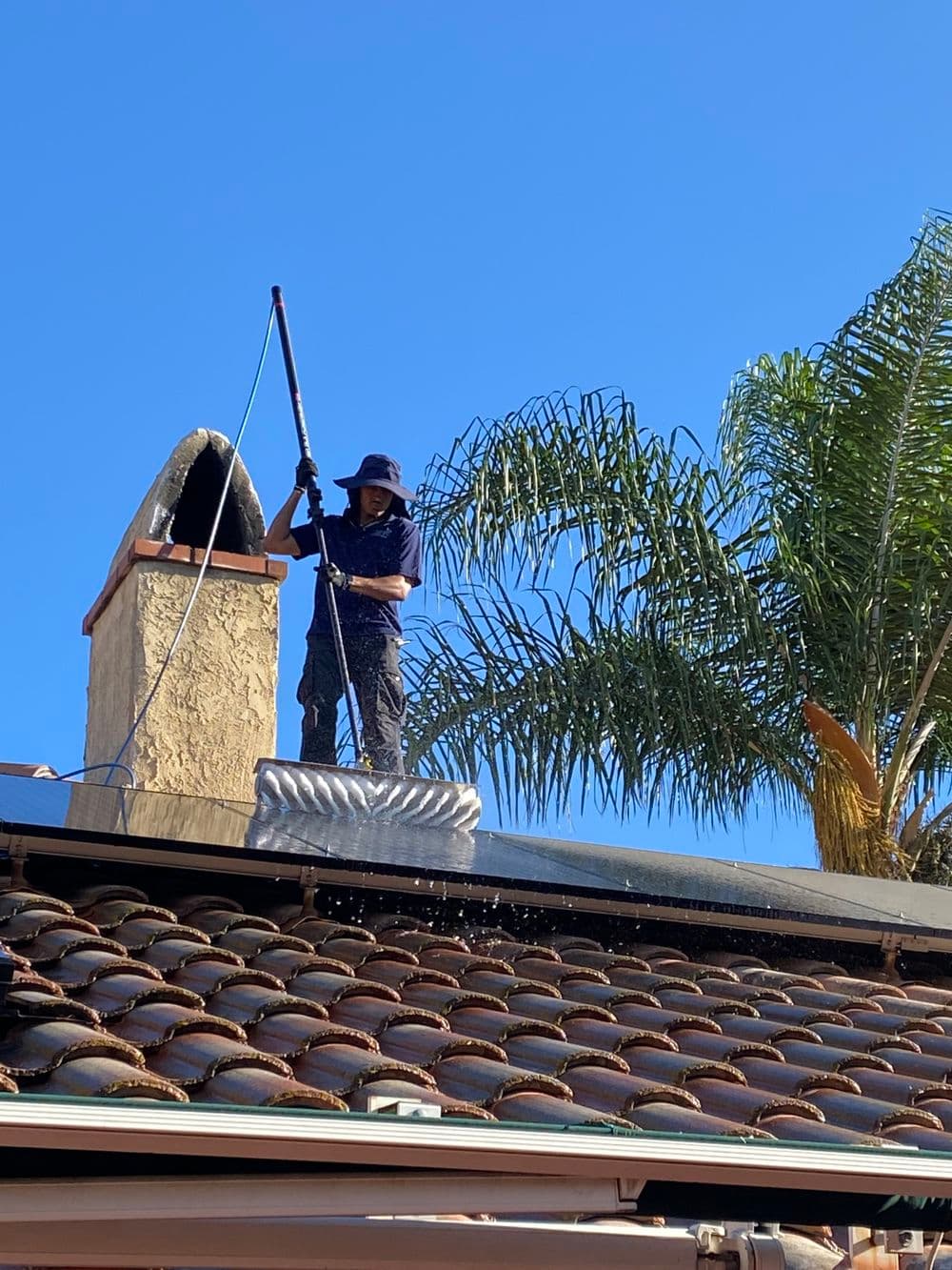 Worker cleaning roof with pressure washer under clear blue sky and palm trees.