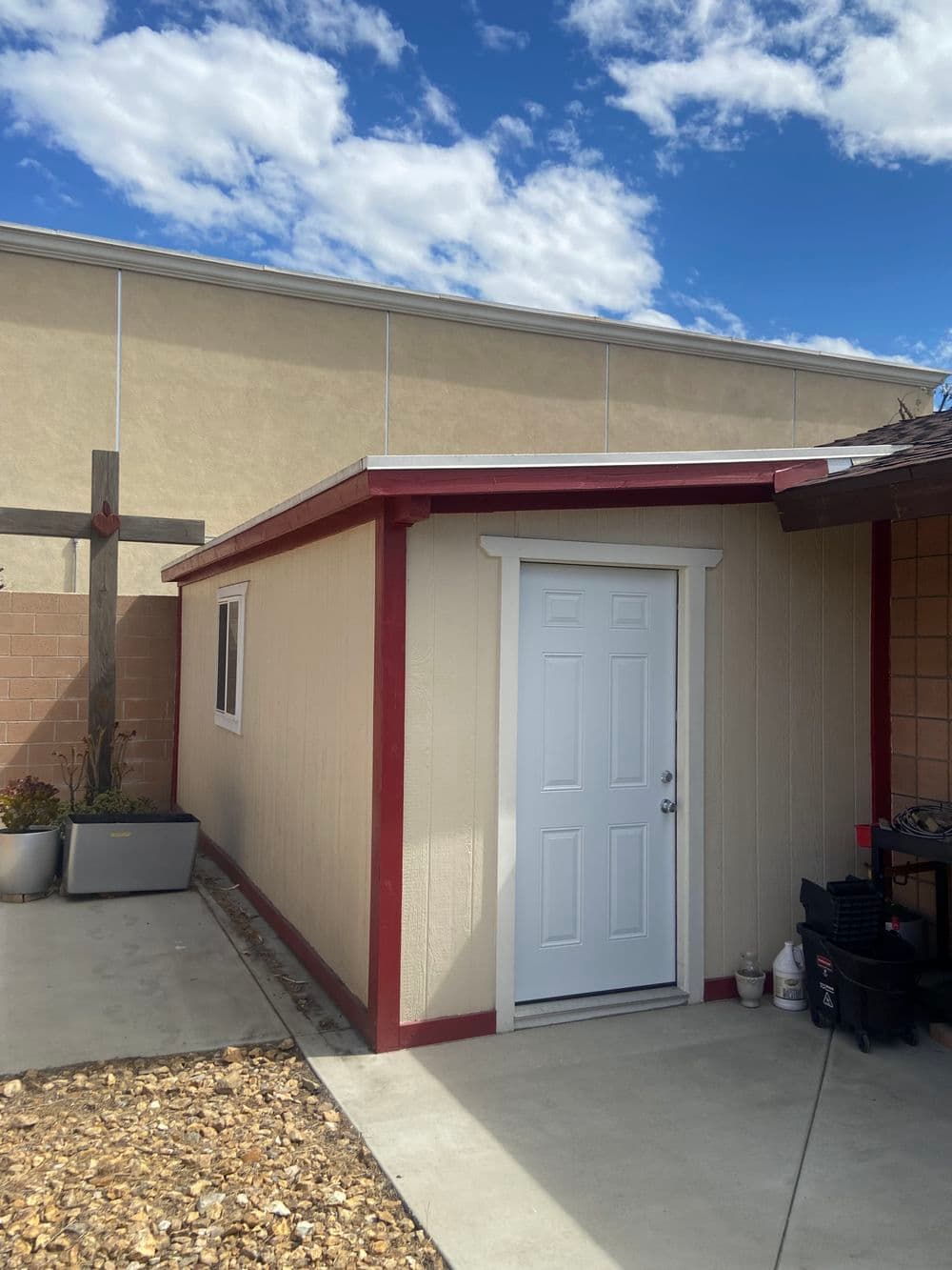 Small storage shed with white door and red trim against a blue sky and building backdrop.