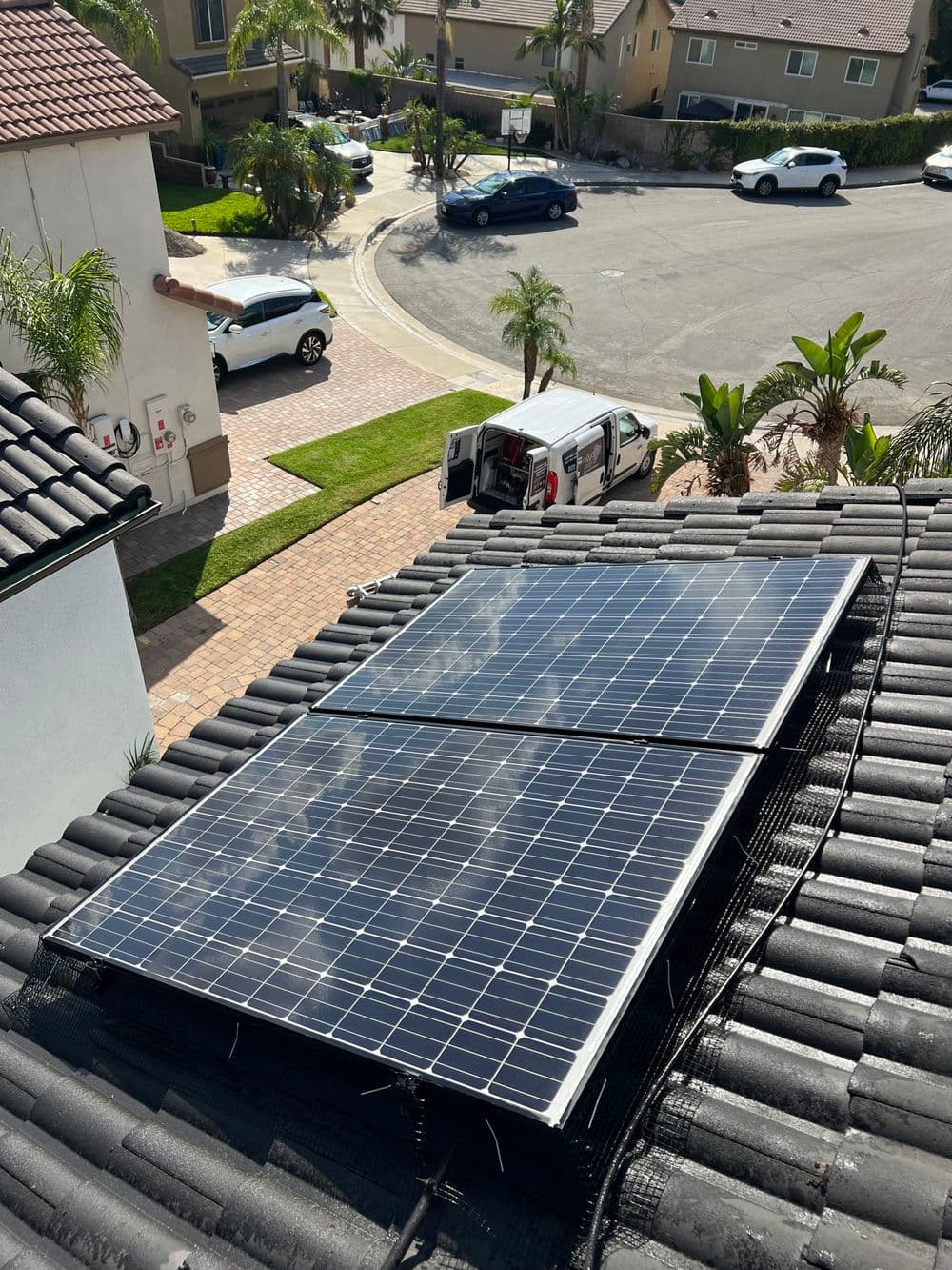 Solar panels installed on a rooftop with palm trees and vehicles in a suburban neighborhood.