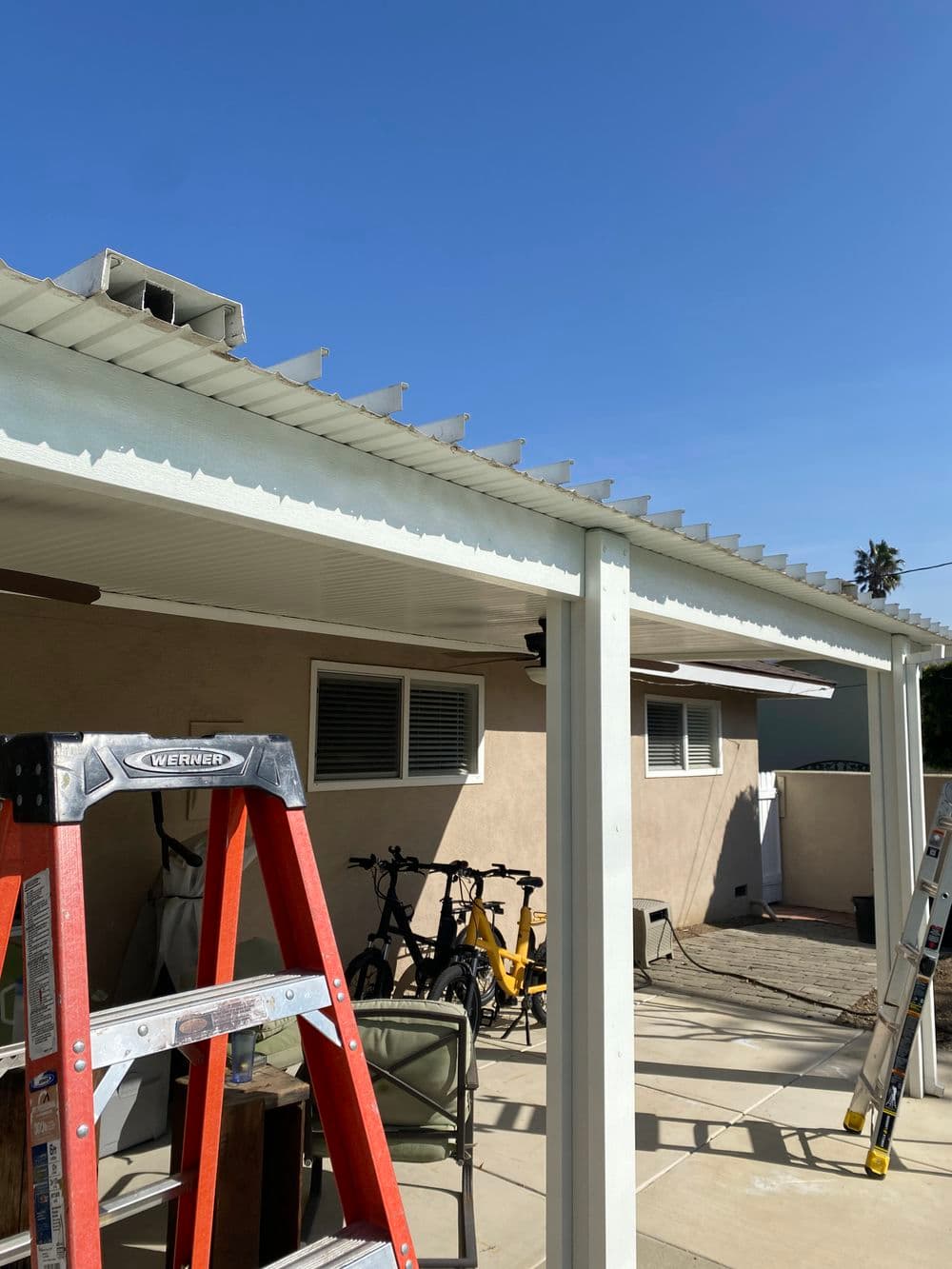 Ladder in backyard under patio cover with blue sky and bicycles in the background.