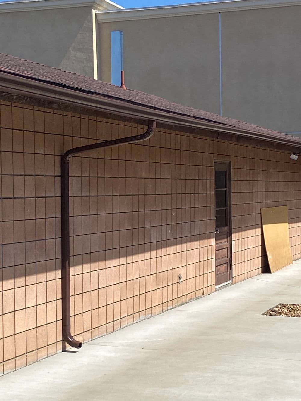Drainage pipe running down the brick wall of a building with a door and wooden panel nearby.