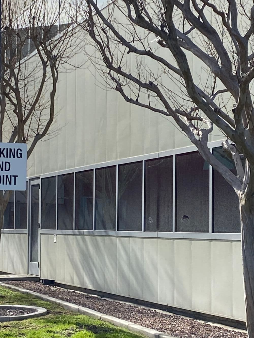 Building exterior with large windows and barren trees in the foreground, parking sign nearby.