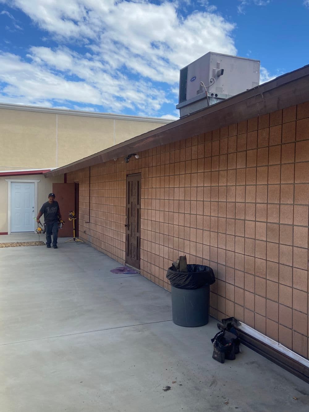 Worker near exterior of building with air conditioning unit and trash can on concrete surface.