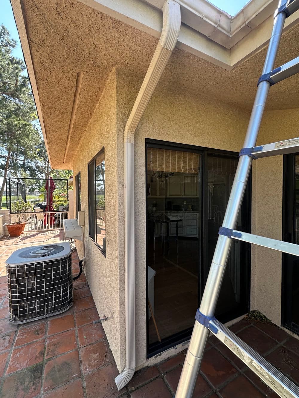 Ladder next to a house with white guttering and air conditioning unit on a sunny day.