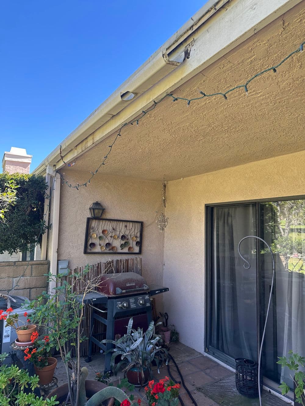 Patio area featuring a grill, plants, and decorative lights against a clear blue sky.